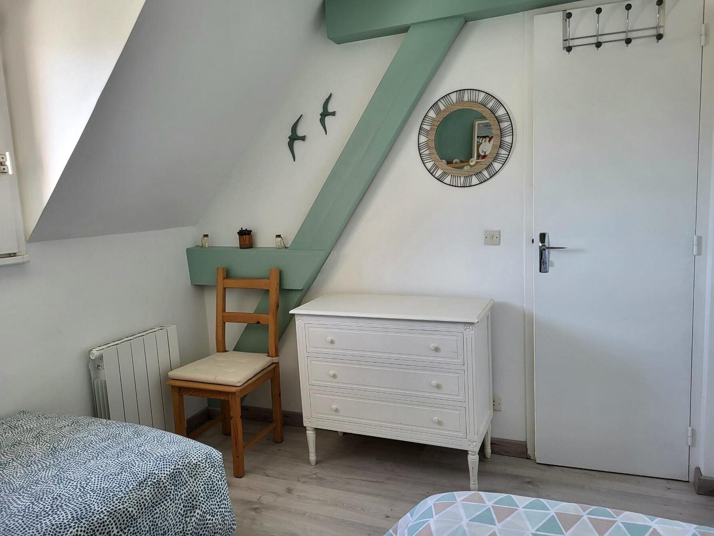 Une chambre avec un plafond en pente, une chaise en bois, une commode blanche, une porte avec un miroir et un radiateur.