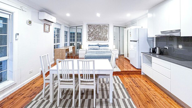 Une cuisine moderne avec des meubles blancs, un coin repas agrémenté d’un tapis à rayures et une chambre avec un lit et une grande fenêtre.