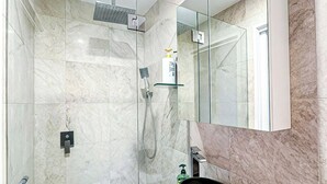 Une salle de bain moderne avec une douche à parois de verre, un mur recouvert de carreaux de marbre, un meuble-lavabo blanc doté d’un lavabo rond et un grand miroir.