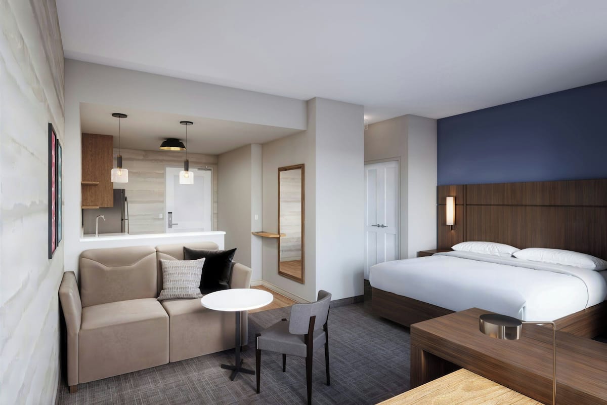 A hotel room with a bed, a sofa, a table, a chair, a small kitchen area, and a bathroom.