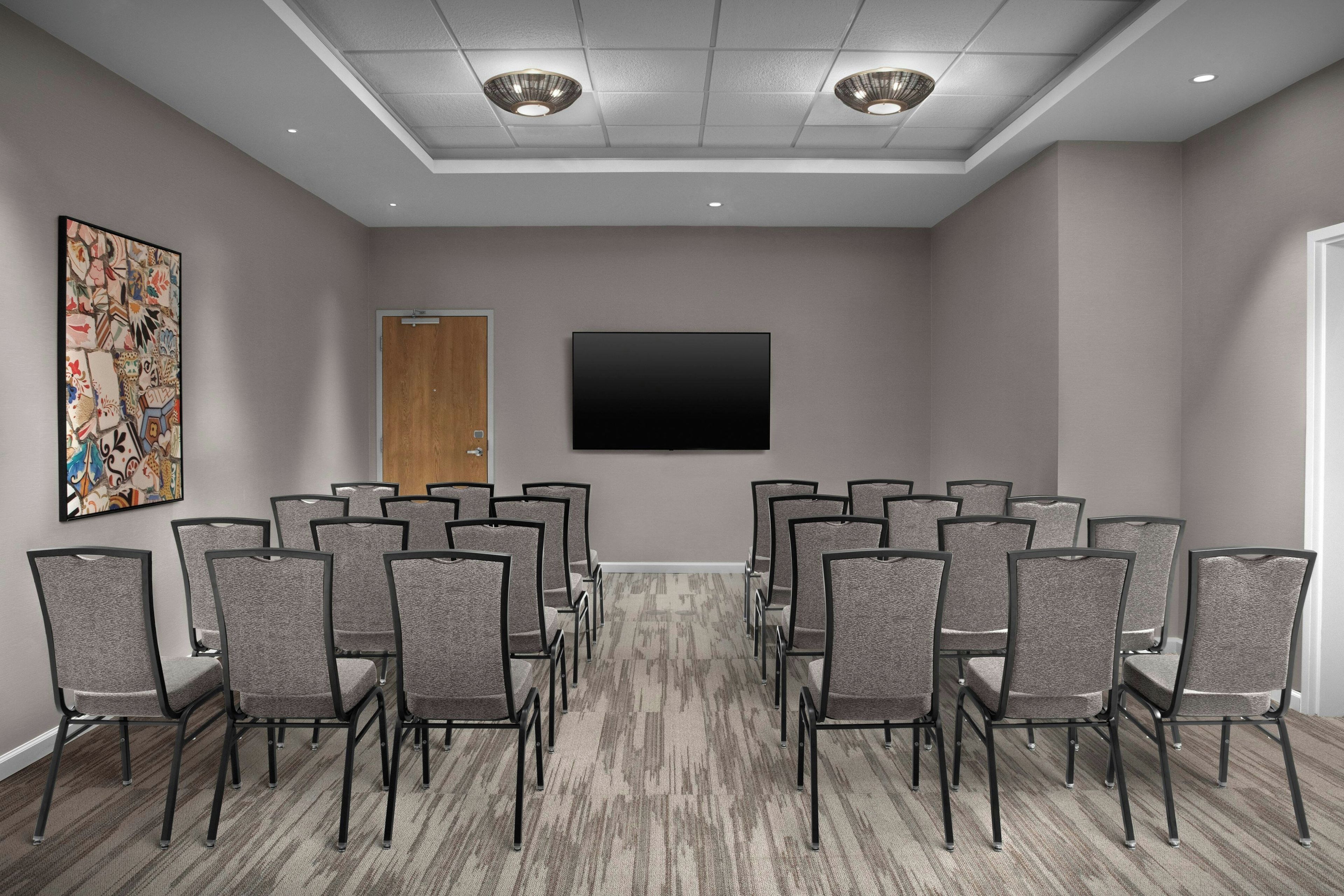 A conference room with a large flat-screen TV, multiple rows of chairs, a carpeted floor, and a door.