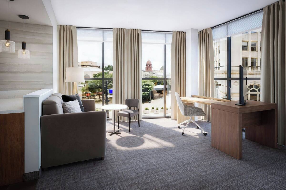 A modern hotel room with a sofa, desk, and chairs, large windows with curtains, and a view of a cityscape.