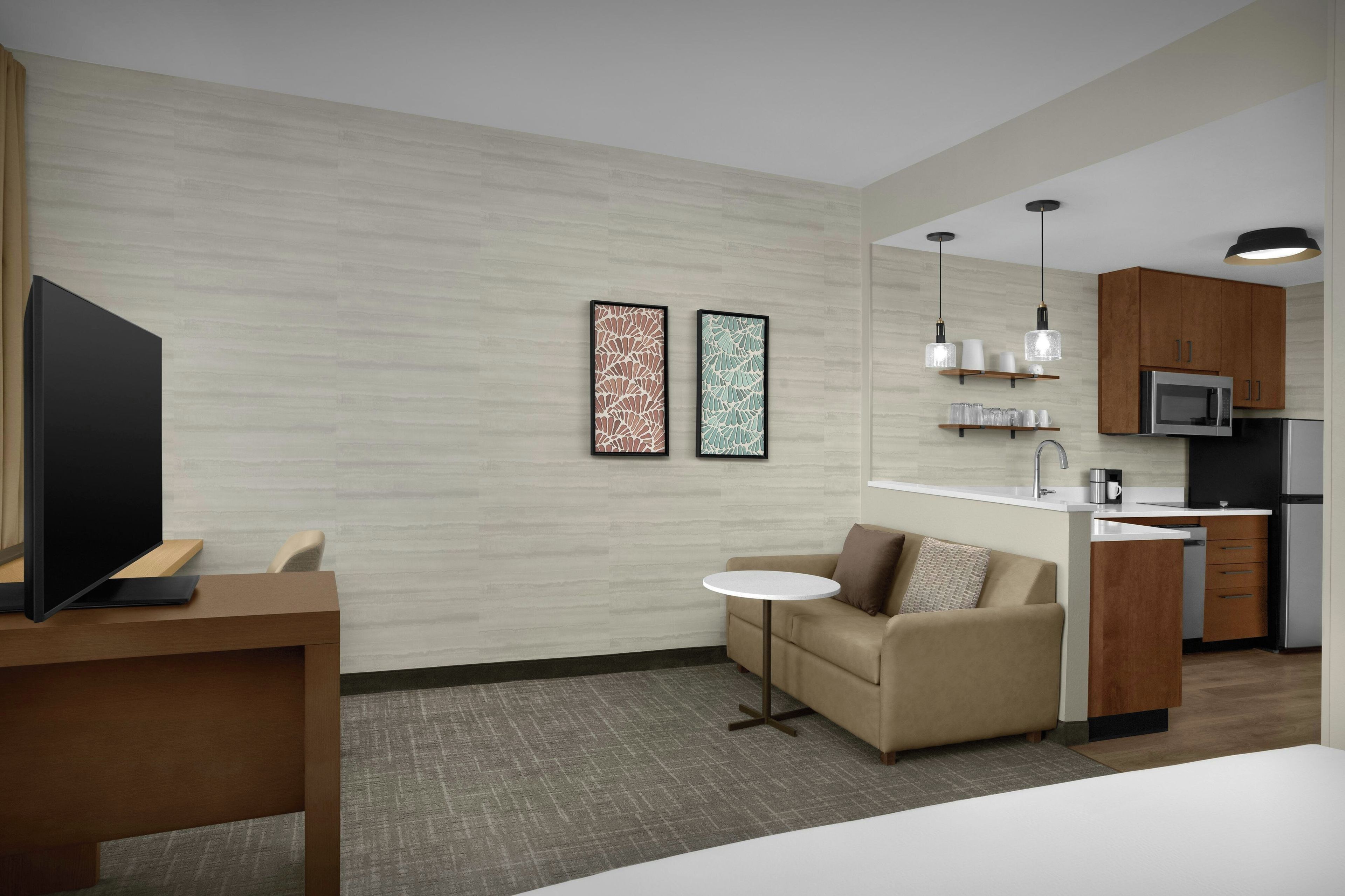 A modern hotel room with a flat-screen TV, a sofa, a small round table, a kitchenette with a microwave and refrigerator, and two framed artworks on the wall.