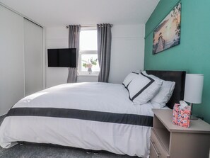 Ein Hotelzimmer mit Bett, Flachbildfernseher, Fenster mit Vorhängen, einem Bild an der Wand und einem Nachttisch mit Lampe und Taschentuchbox.