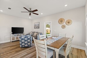 A dining area with a wooden table, white chairs, a ceiling fan, a television on a white cabinet, and a door leading outside. - Amazing 3-bedroom townhome steps from beach and many attractions PCB (Panama City Beach)