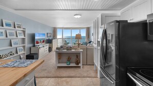 Uma cozinha moderna com geladeira preta, uma área de refeições com mesa de madeira e vista para o mar através de grandes janelas.