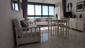 Ein Wohnzimmer mit einem weißen Sofa, einem Esstisch mit Stühlen, einem Fernseher und einem Fenster mit Vorhängen.