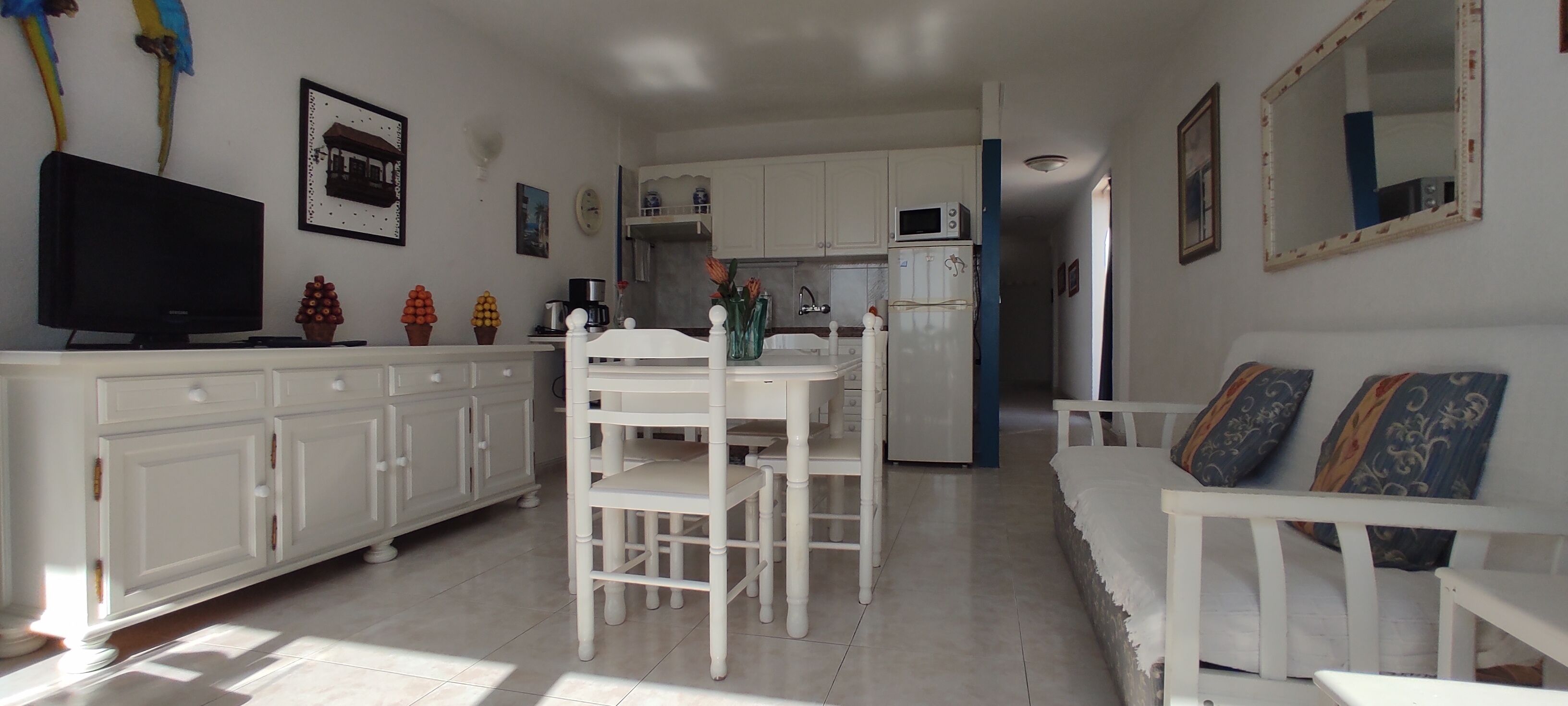 Een ruimtelijk ingerichte leefruimte met kitchenette, een eethoek met witte meubels en een zithoek met gestreepte kussens.
