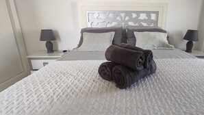 Ein ordentlich gemachtes Bett mit aufgerollten Handtüchern, einer weißen, strukturierten Tagesdecke und einem Kopfteil mit dekorativem Muster.
