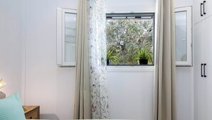 Ein Zimmer mit Fenster, Vorhang, Nachttisch und Bett.