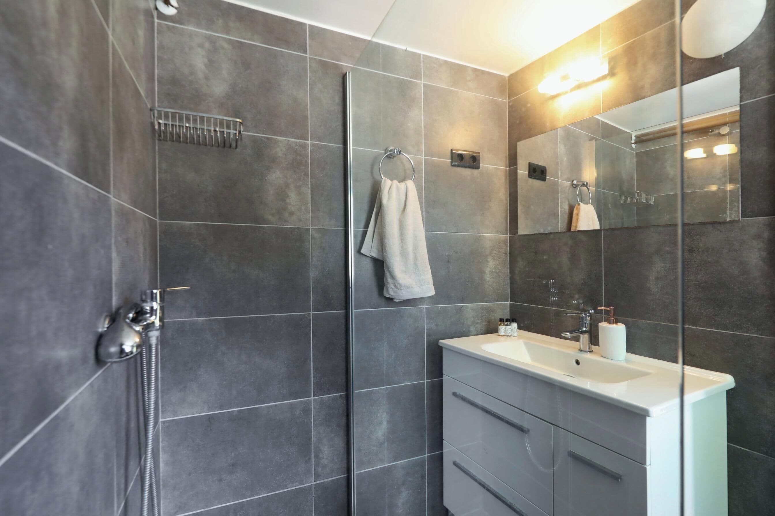 A modern bathroom with grey tiles, a glass shower enclosure, a white sink, and a large mirror.
