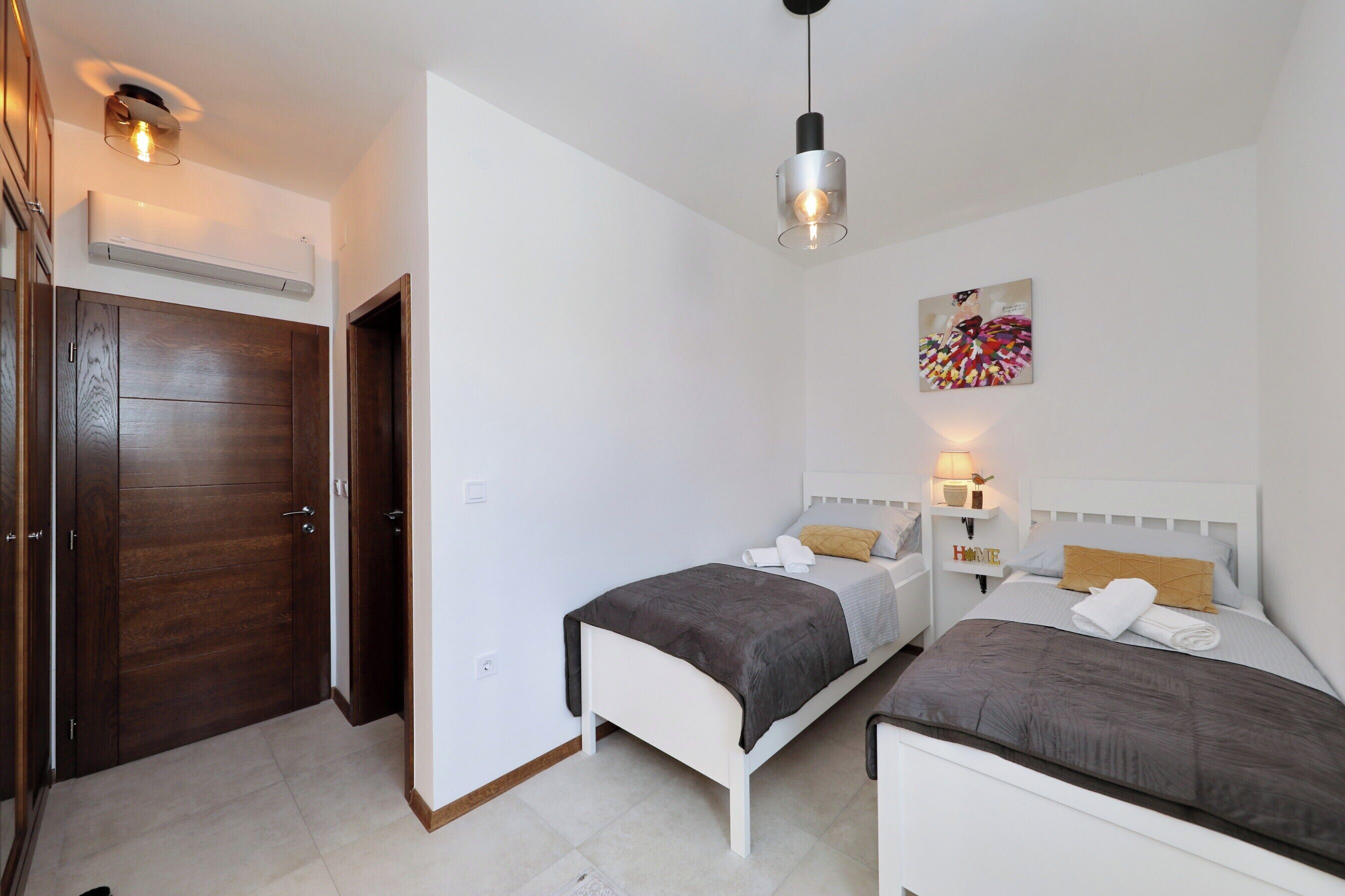 A bedroom with two beds, a wooden wardrobe, an air conditioning unit, a hanging light fixture, and a wall-mounted artwork.