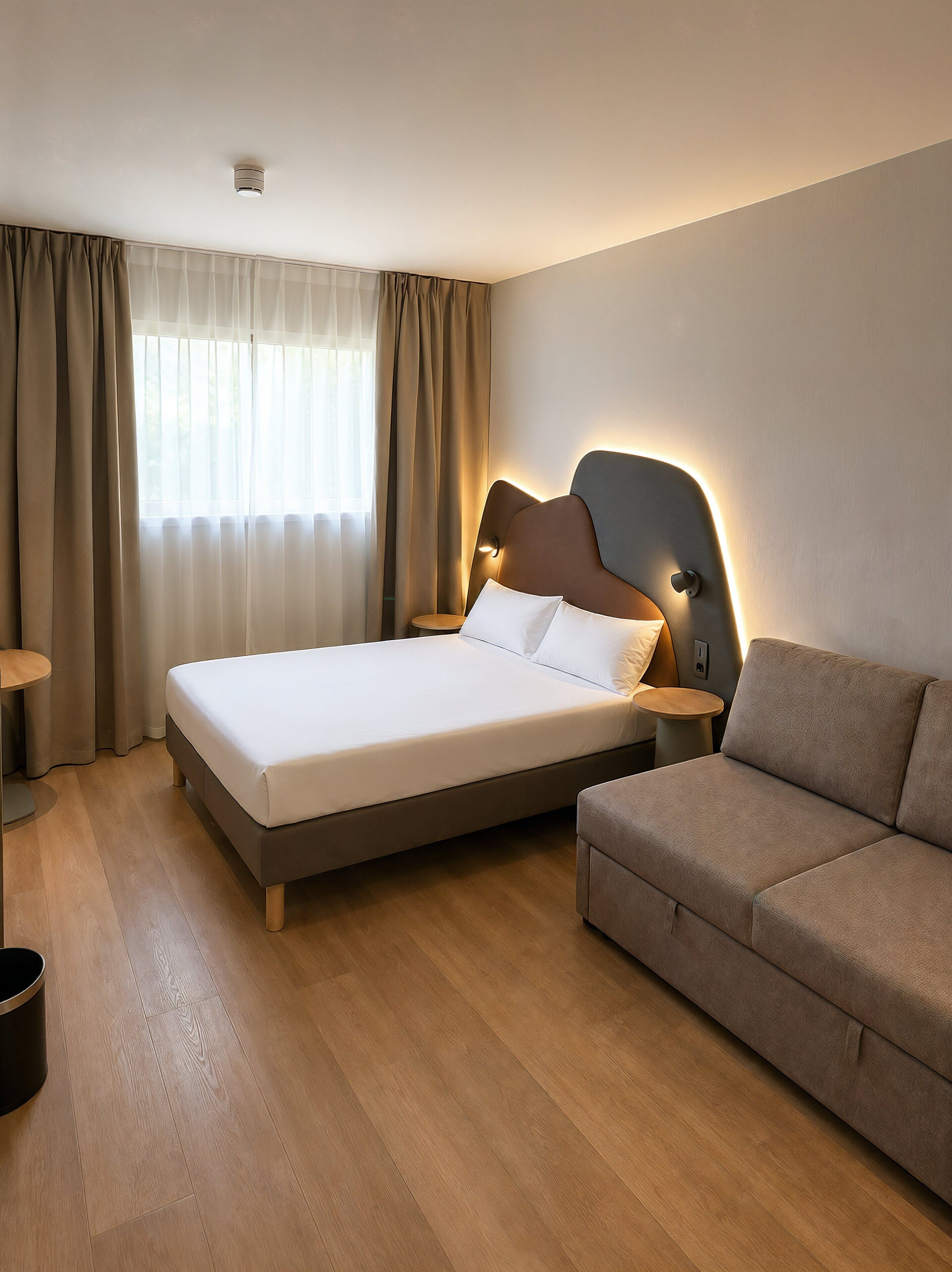 A modern hotel room with a large bed, a sofa, a small table, and a window with curtains.
