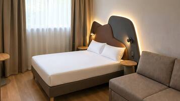 A modern hotel room with a large bed, a sofa, a small table, and a window with curtains.