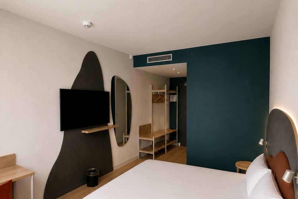 A modern hotel room with a bed, a desk, a TV mounted on the wall, a mirror, and a wardrobe.