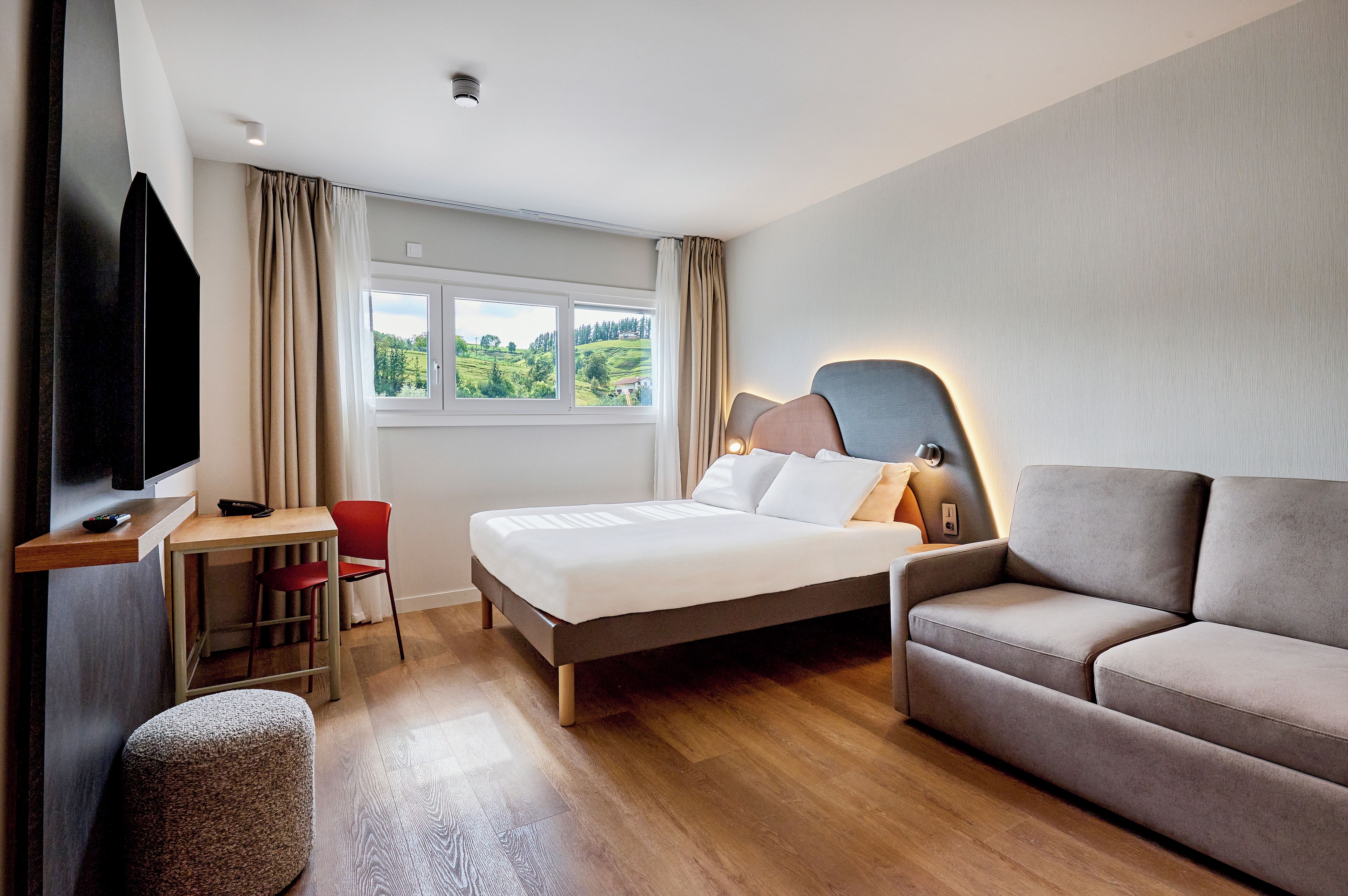 Ein Hotelzimmer mit Bett, einer Couch, einem Schreibtisch mit Stuhl, einem Fernseher und einem Fenster mit Vorhängen.