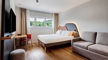 Ein Hotelzimmer mit Bett, einer Couch, einem Schreibtisch mit Stuhl, einem Fernseher und einem Fenster mit Vorhängen.