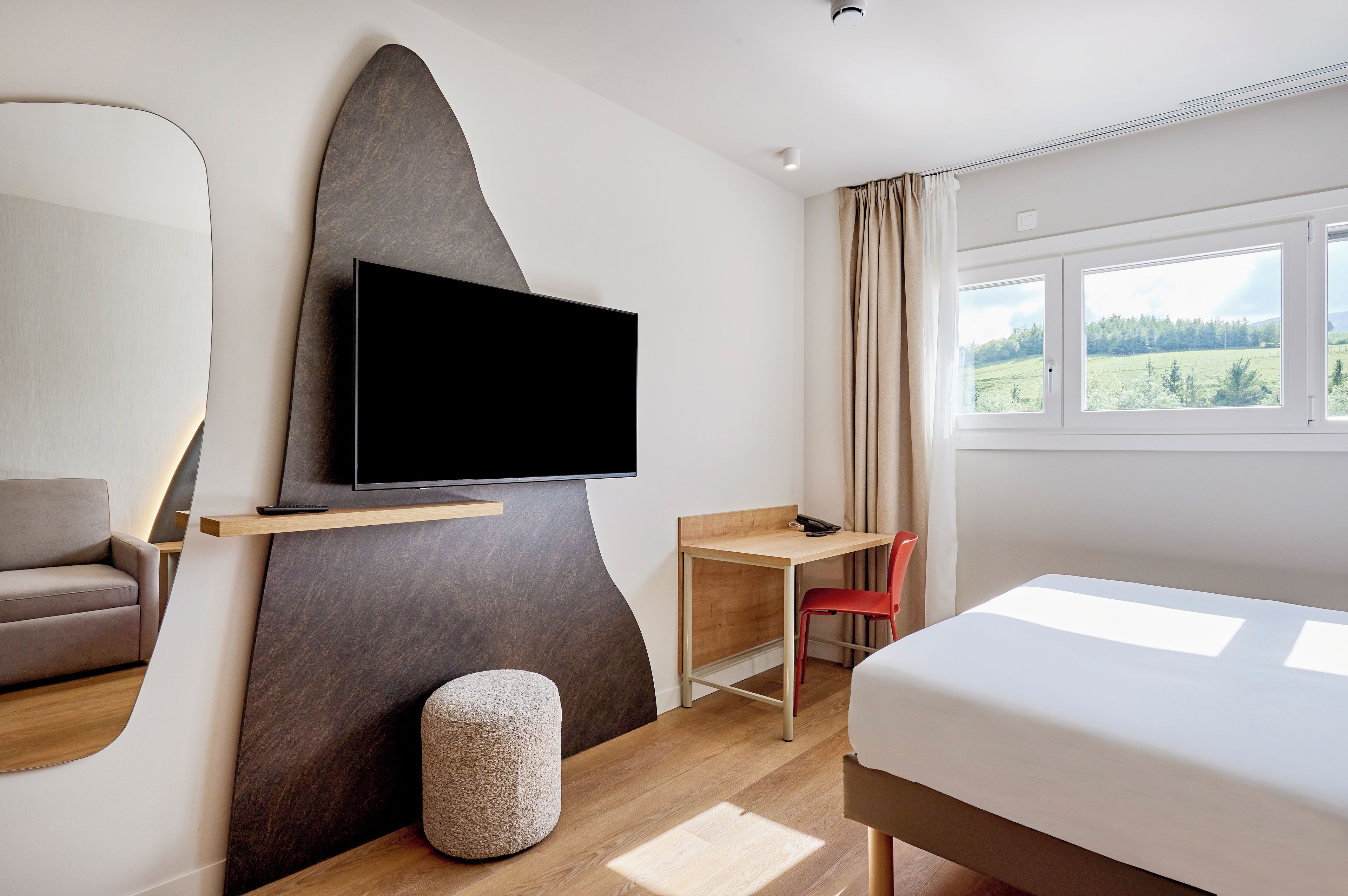 A modern hotel room with a flat-screen TV mounted on a wall, a wooden desk, a red chair, a bed, and a window with a view of greenery.