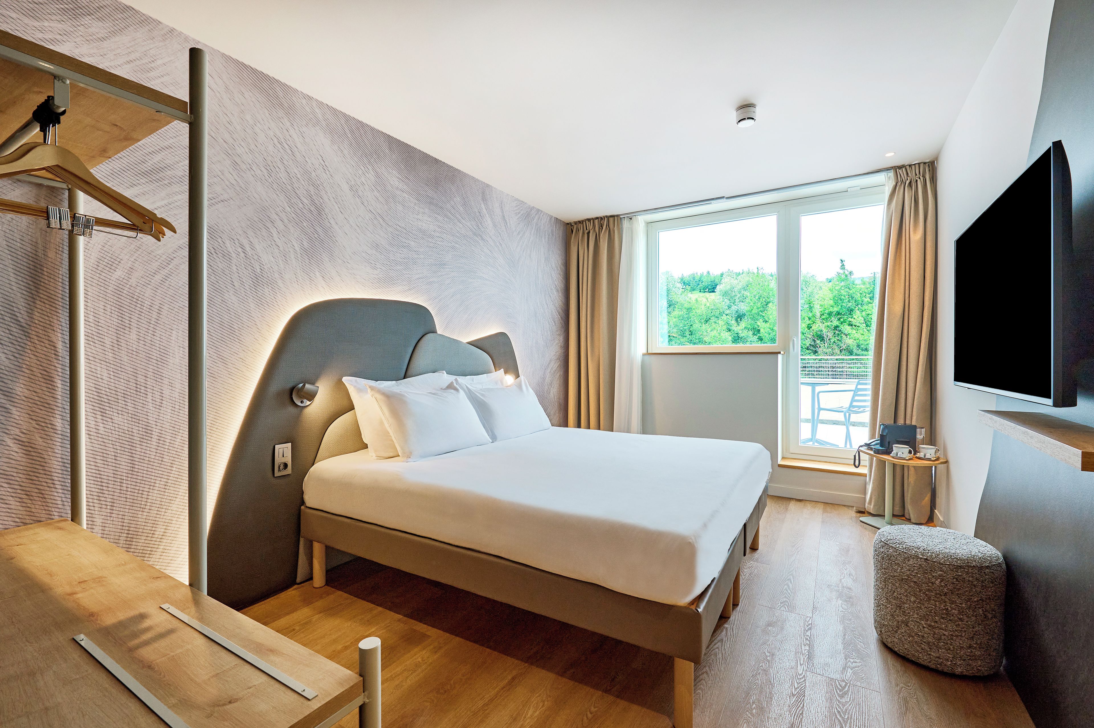 A modern hotel room with a bed, a television, a window with curtains, and a wooden floor.