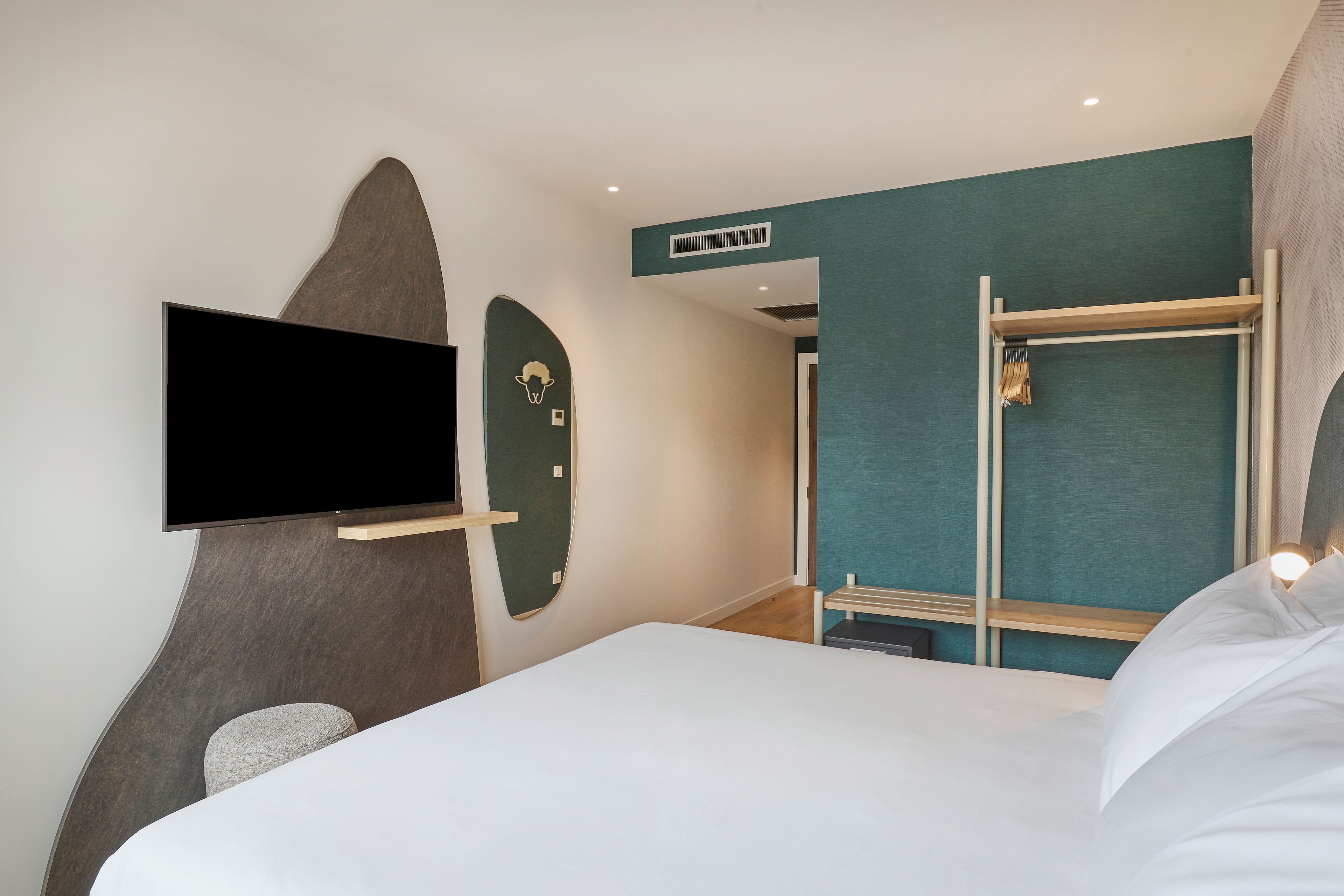 A hotel room with a bed, a flat-screen TV mounted on the wall, a wooden shelf, and a wardrobe with a mirror.