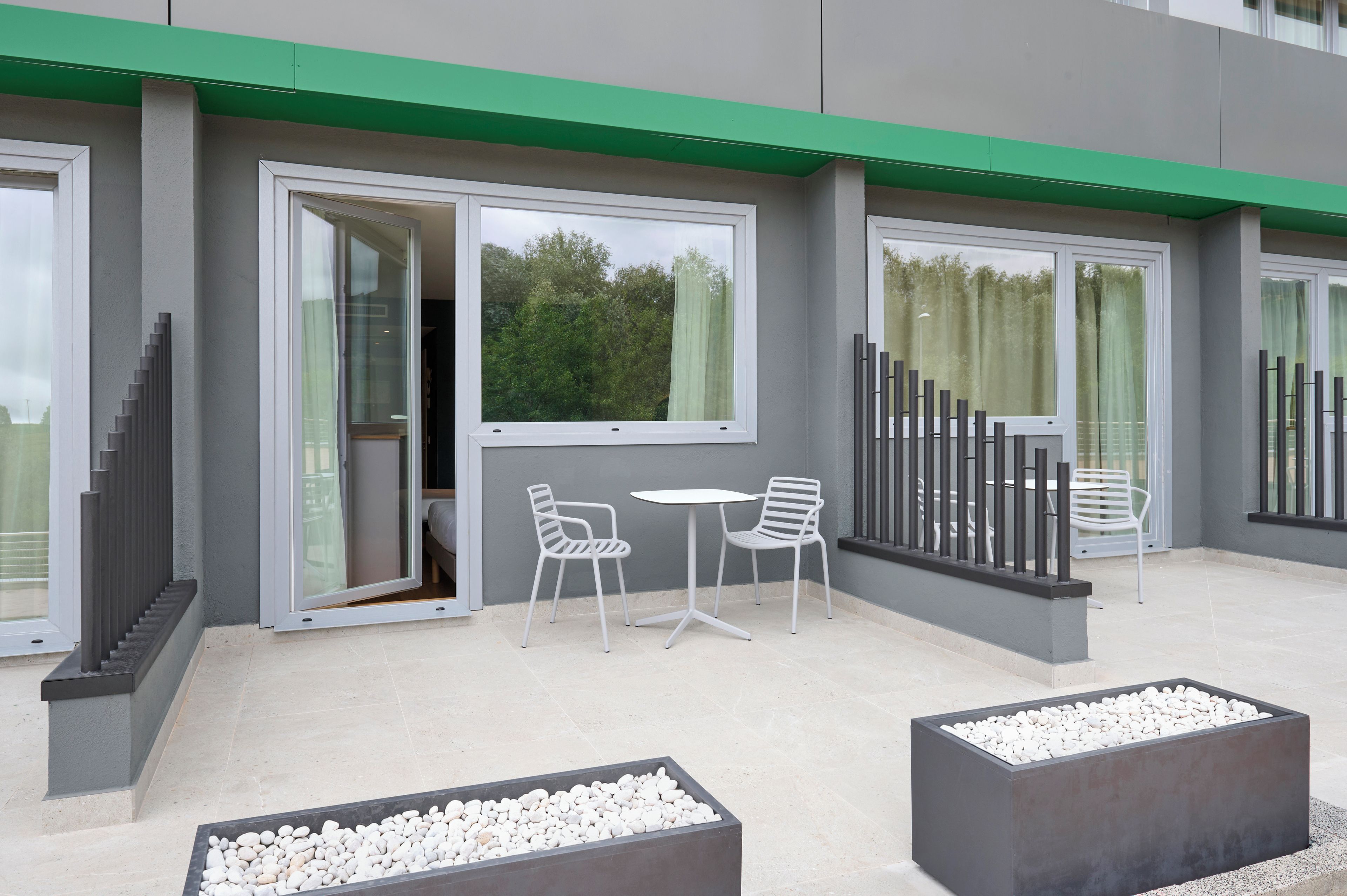 A modern outdoor seating area with two white chairs and a table, surrounded by grey walls and large windows.