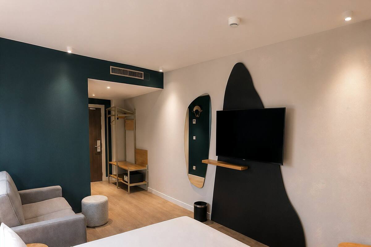 A modern hotel room with a bed, seating area, TV, and a view of the hallway.