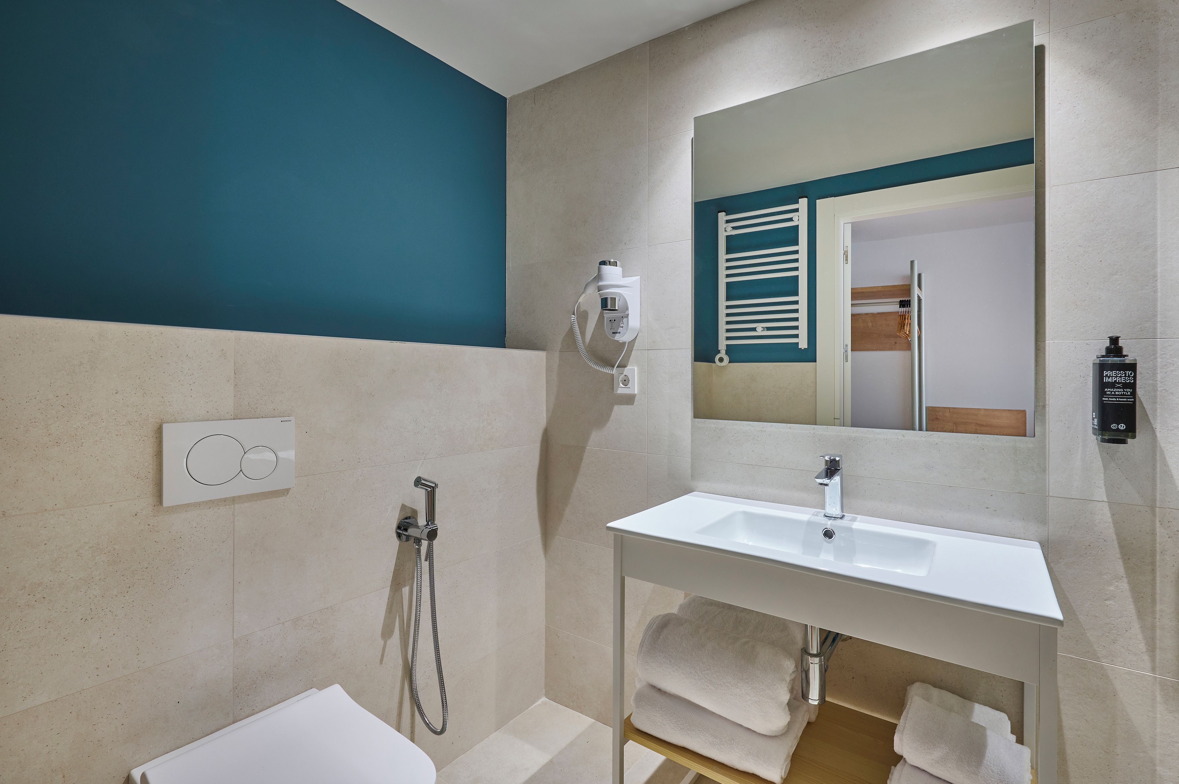 Un baño moderno con un lavamanos blanco, un cabezal de ducha manual, un perchero y un espejo con reflejo de una puerta de madera.