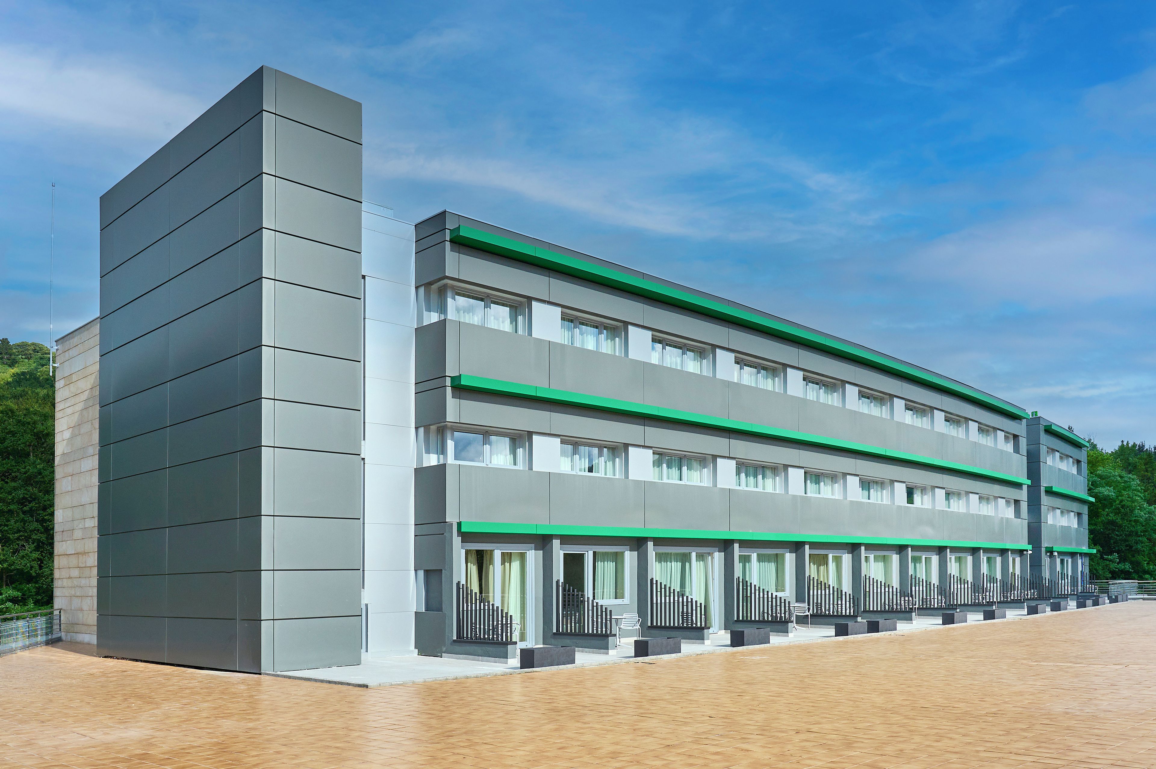 Un edificio moderno con techo plano, múltiples ventanales y un detalle verde en los pisos superiores.