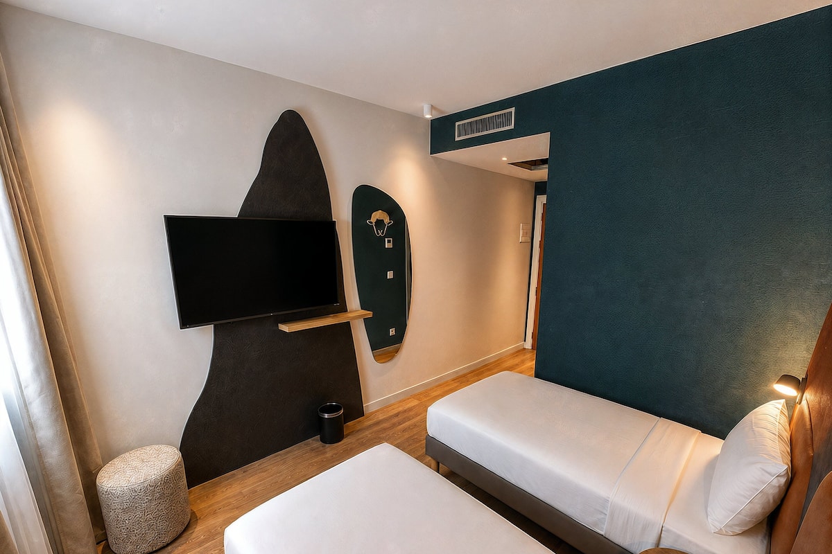 A hotel room with a bed, a flat-screen TV mounted on the wall, a bedside table, and a small round ottoman.