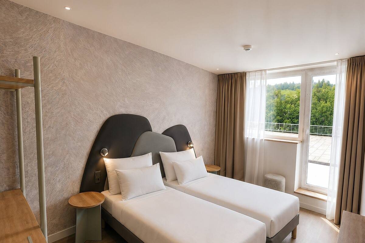 A hotel room with two beds, a wooden desk, a small round table, and a window with a view of greenery.