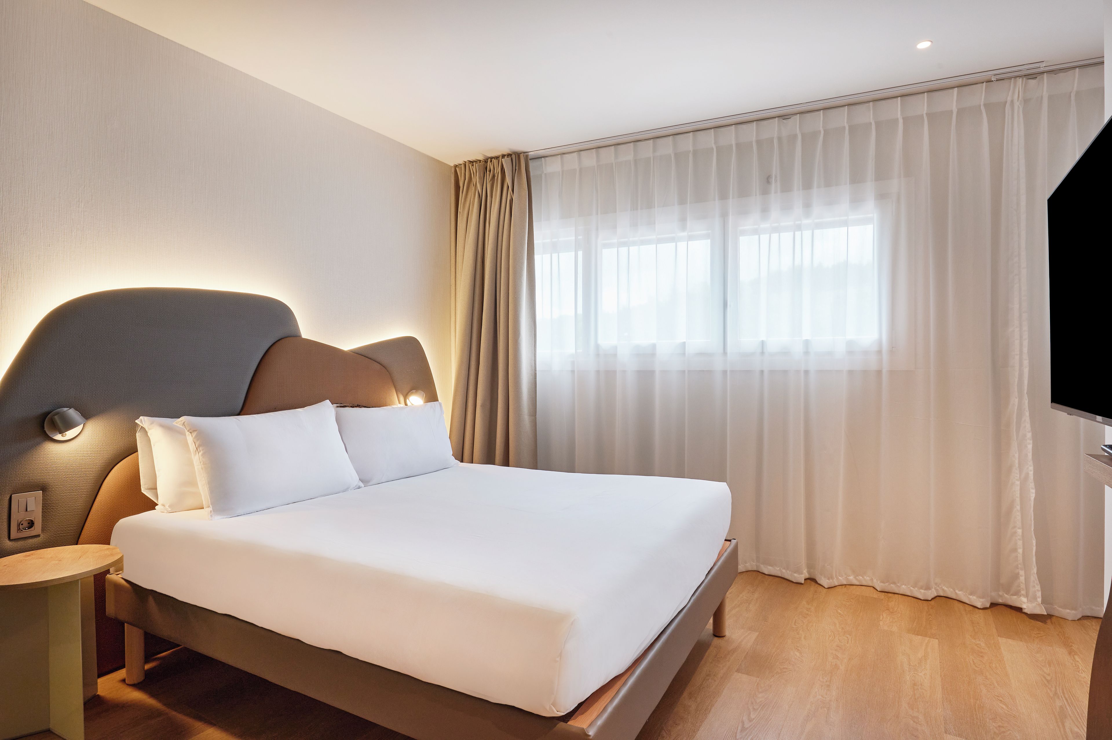 Habitació d'hotel amb llit, taulell, làmpada de paret, finestra amb cortines lleugeres i televisió de pantalla plana.