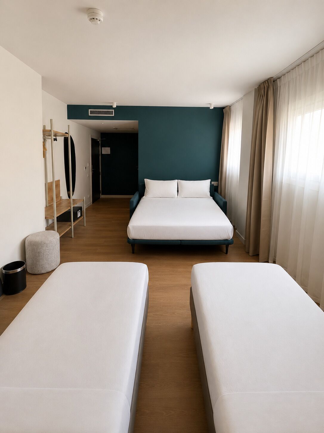 Une chambre d’hôtel avec deux lits, un bureau, une chaise, un petit tabouret, une poubelle et une porte donnant sur une autre pièce.