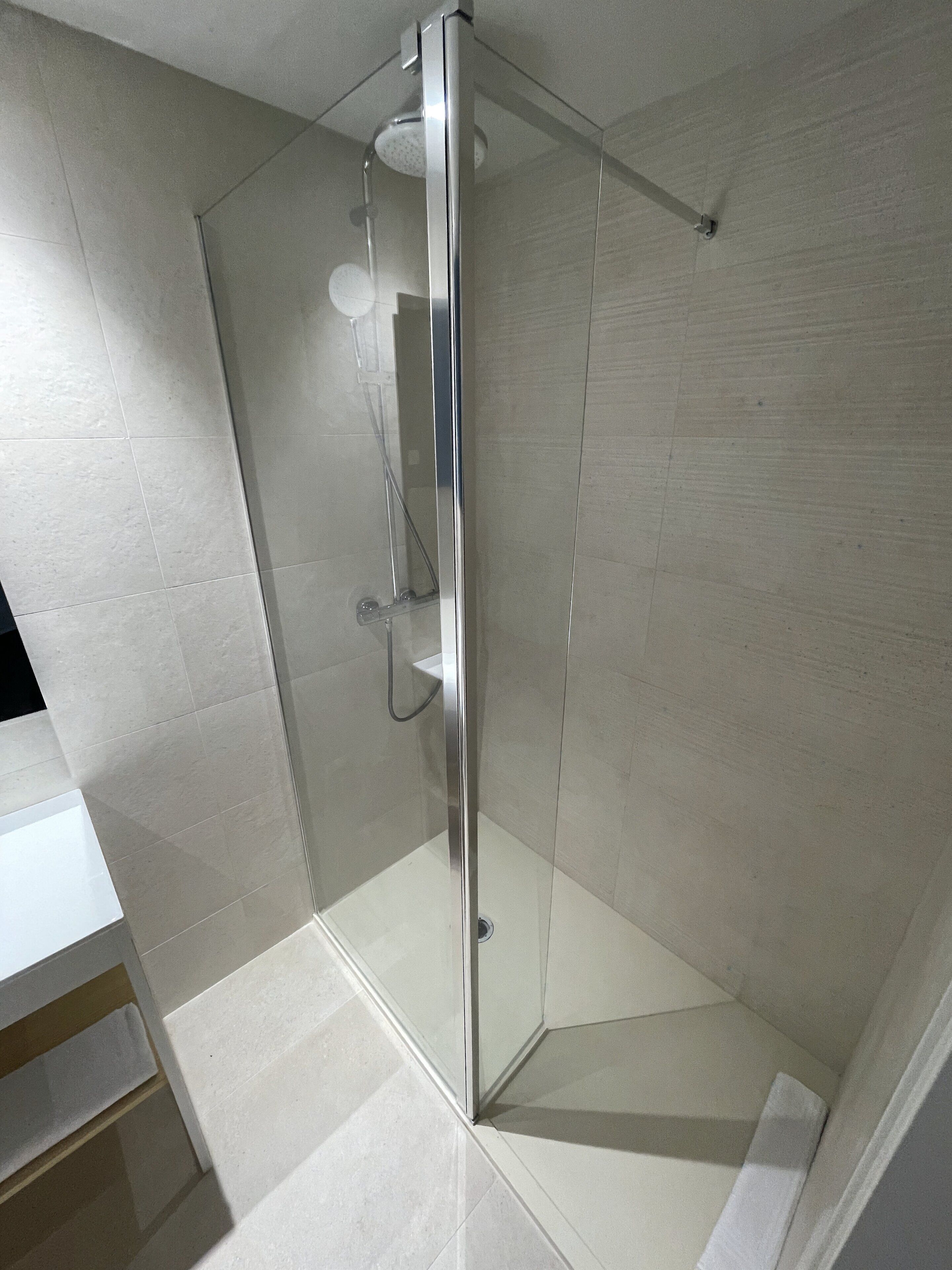 Ducha con mampara de vidrio, estructura de cromo, piso de baldosas blancas y desagüe visible.