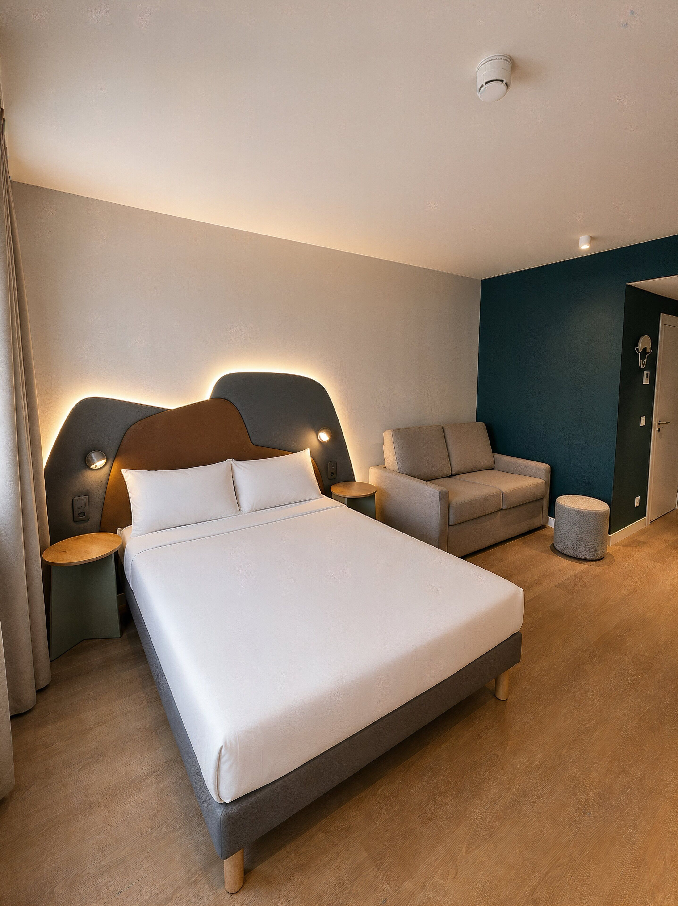 A modern hotel room with a large bed, a sofa, a small table, and a round ottoman.