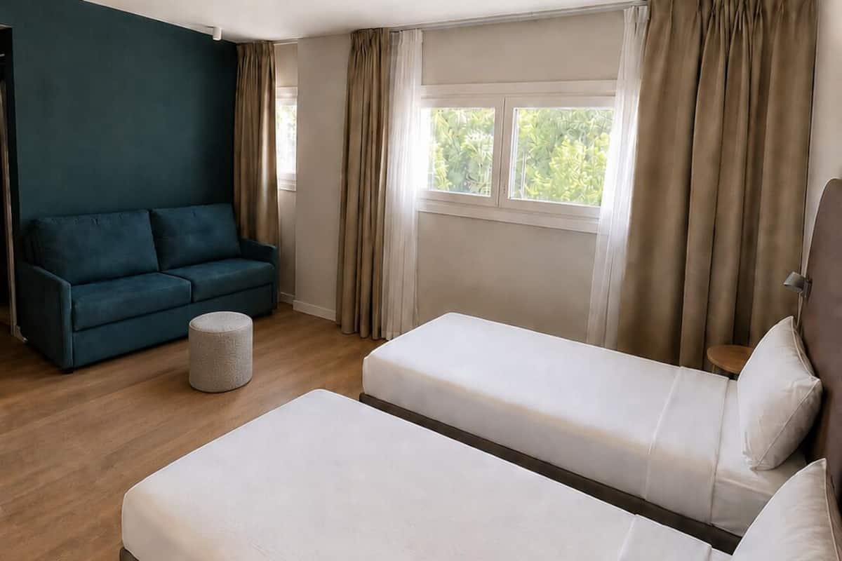 A modern hotel room with a large bed, a sofa, a small round ottoman, and a window with curtains.