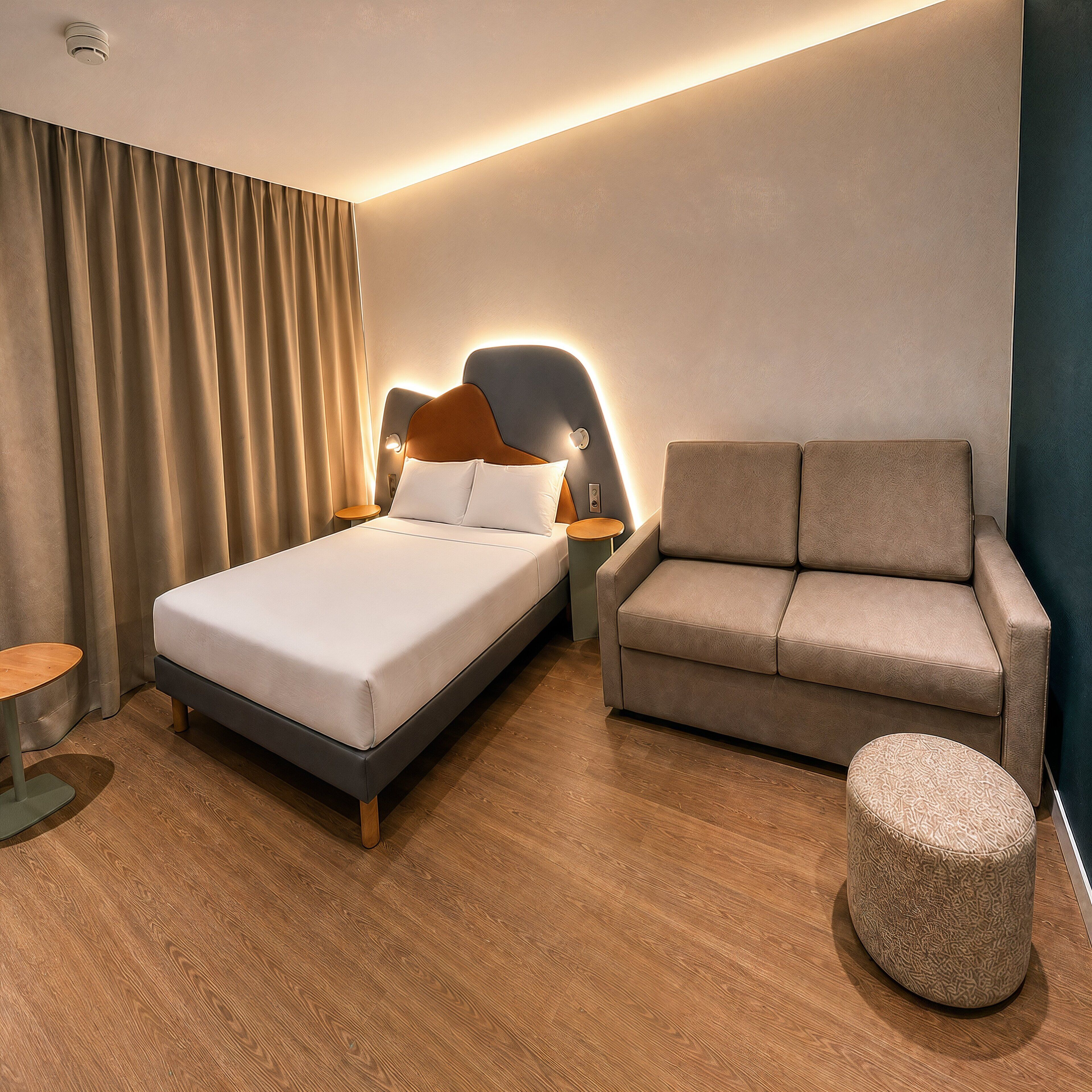 Une chambre d’hôtel comprenant un lit, un canapé, un pouf rond et une table de chevet avec une lampe.