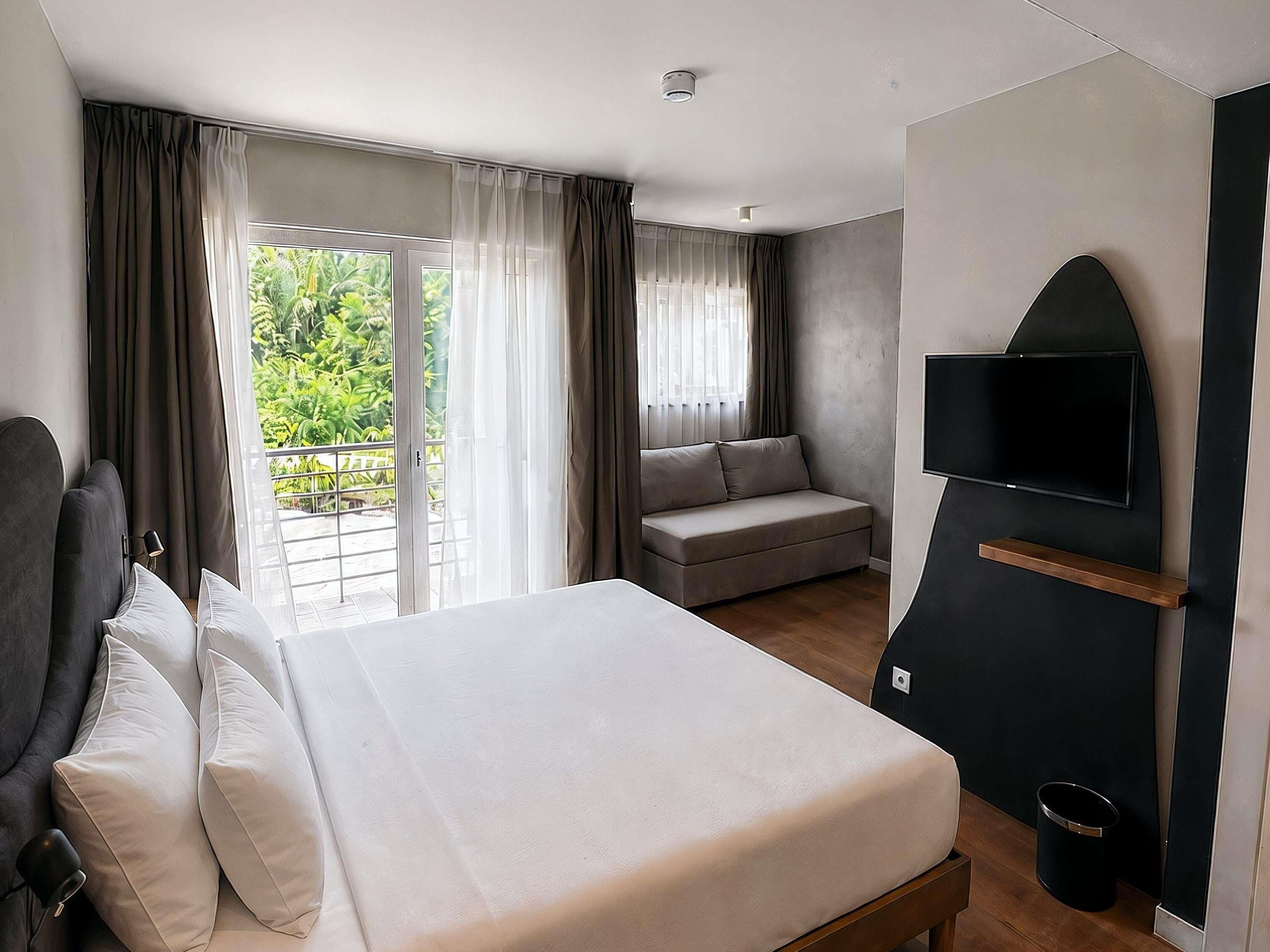 Ein Hotelzimmer mit Bett, einer Couch, einem Fernseher und einem Balkon mit Vorhängen.