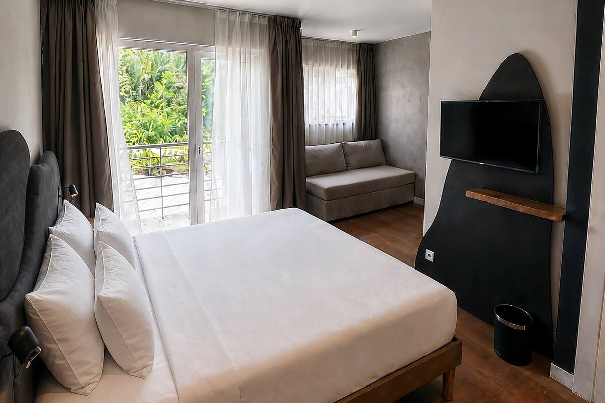 A modern hotel room with a large bed, a sofa, a TV mounted on the wall, and a balcony with curtains.