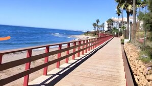 A wooden boardwalk with red railings, leading to a beach with palm trees and a building in the background. - Algaida Beach Send Litoral Studio with Sea View, Adults Only (Calahonda)