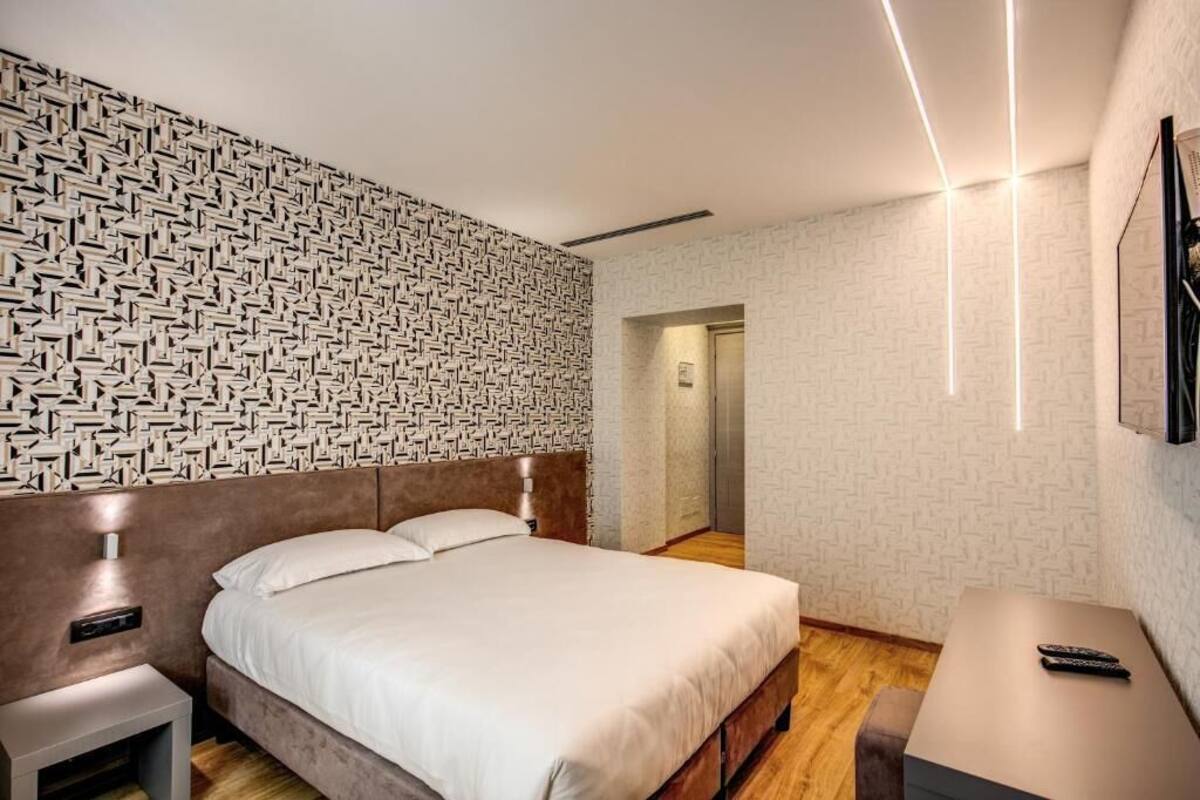 A modern hotel room with a patterned wall, a bed, a bedside table, a nightstand, a small table, and a remote control on the table.
