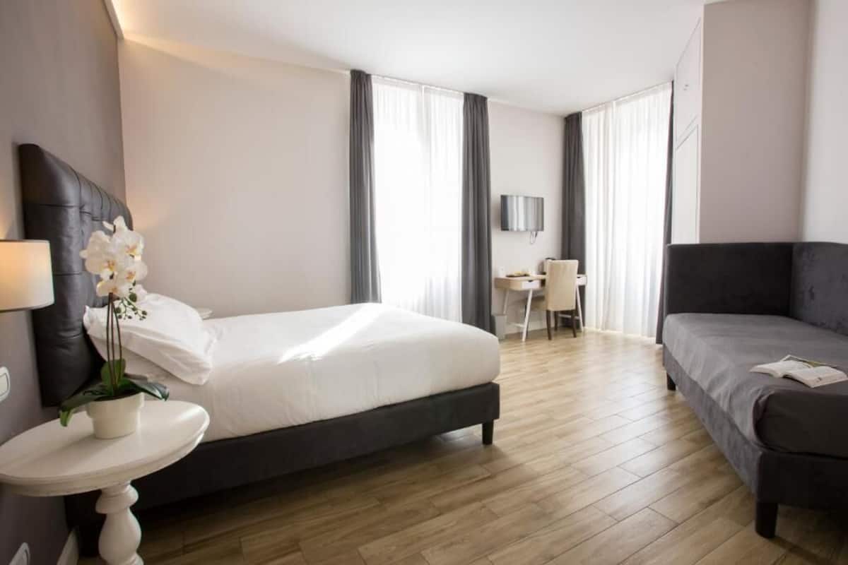 A hotel room with a bed, a sofa, a small table with a lamp, a desk, and a TV.