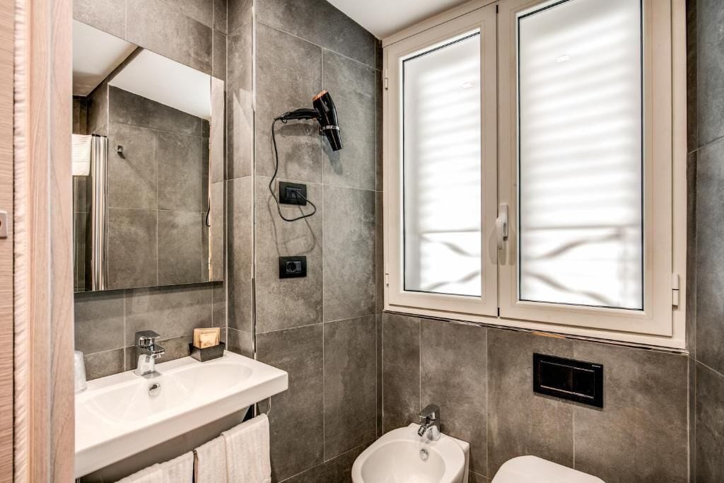 A modern bathroom with a large mirror, two sinks, a bidet, and a hairdryer mounted on the wall.