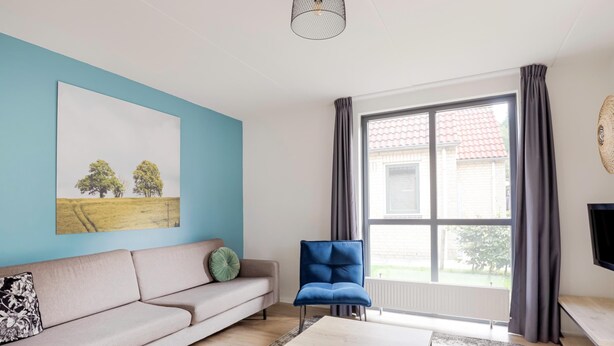 A modern living room with a sofa, a coffee table, a blue chair, a large window with curtains, and a painting on the wall. - Tranquil Ewijk Escape for 6 (Ewijk)