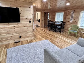 A wooden interior with a flat-screen TV, a dining table with chairs, a green chair, and a grey sofa. - Junction: Lake Life & Village Charm – Sleeps 4 (Walloon Lake)