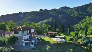Una casa de dos pisos con balcón, rodeada de abundante vegetación y colinas.