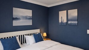 A bedroom with a white bed, two blue pillows, a white headboard, and two landscape paintings on the wall.