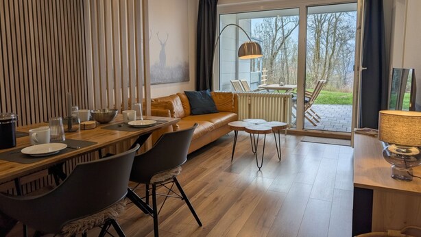 A modern living room with a wooden dining table, leather sofa, and a large sliding glass door leading to an outdoor area.
