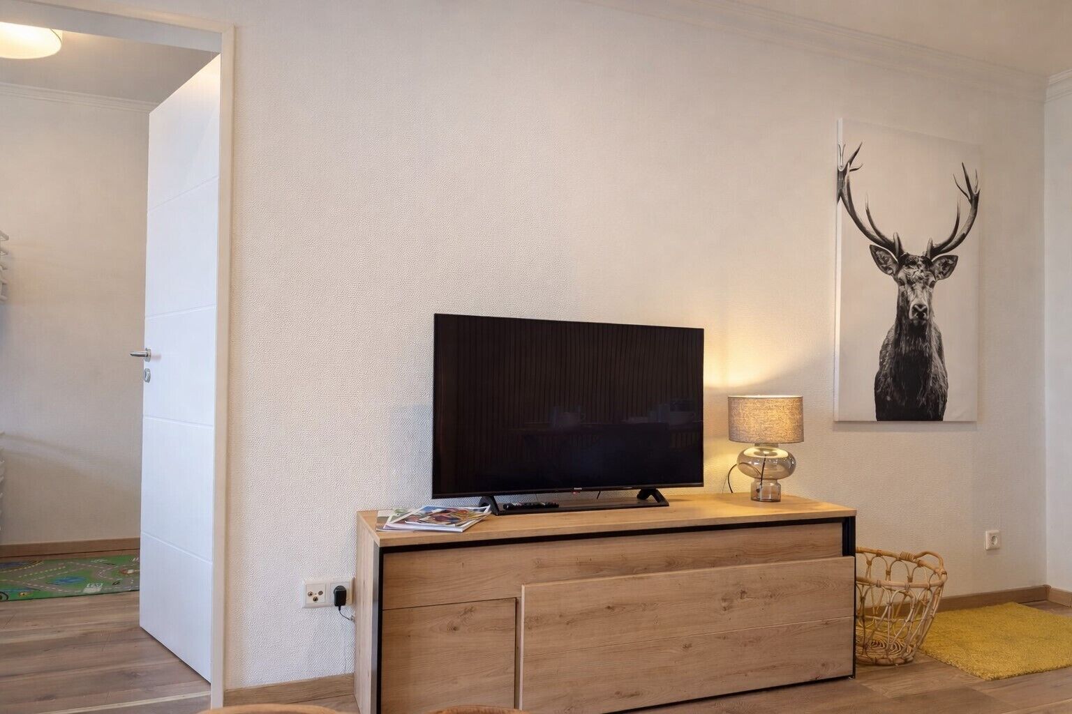 A room with a TV on a wooden cabinet, a lamp, a wall art of a deer head, and a basket.