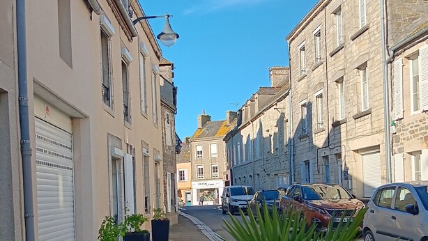 A narrow street lined with stone buildings, parked cars, and potted plants. - Le Picoteu' (Saint-Vaast-la-Hougue)