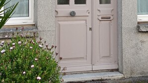 A building with a double door, a window with a number '16' above it, and a small plant with flowers in front. - Le Picoteu' (Saint-Vaast-la-Hougue)
